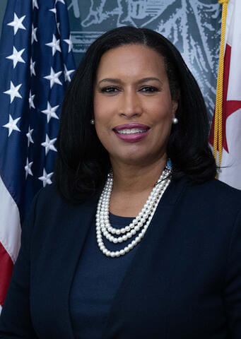 Mayor Muriel Bowser smiling in front of the United States flag and the Washington, DC flag.